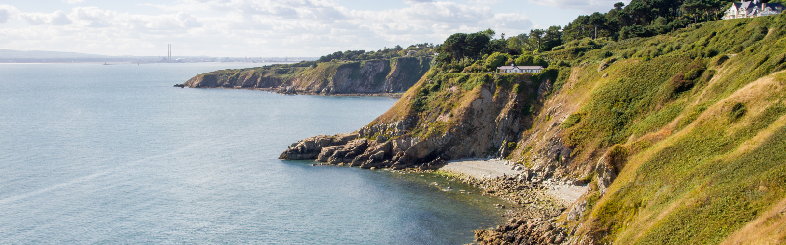 A green cliff with a house very far in the distance touches the edge of a large body of water. 